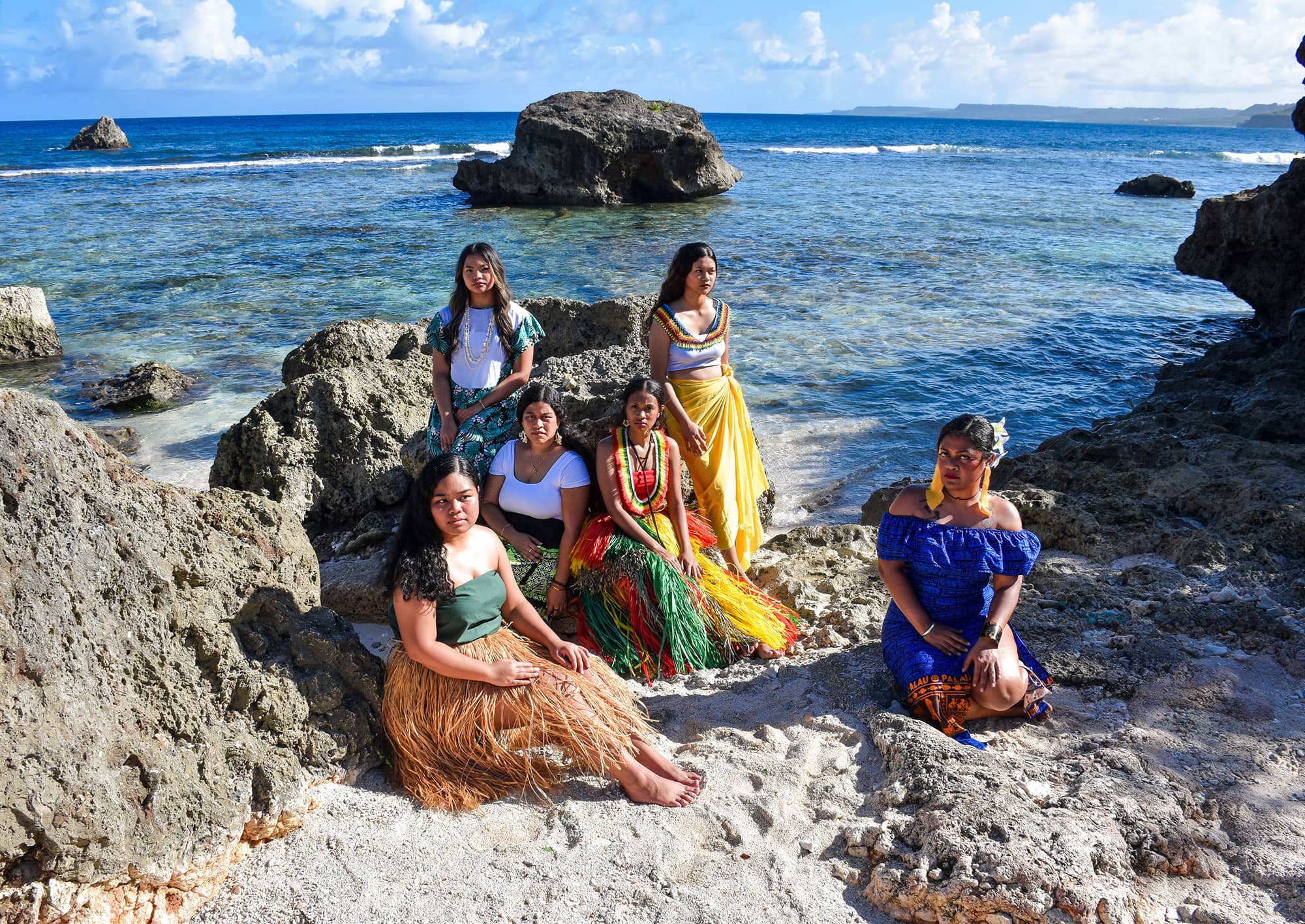 Six women in traditional attire pose on rocky shores with the ocean in the background