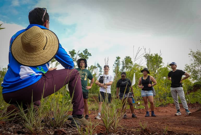 Man sits by plants in red dirt area, talking and facing several other people